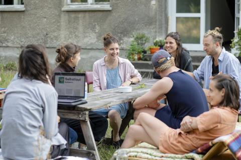 sieben junge Menschen sitzen an einem Tisch vor einem Wohnhaus und reden